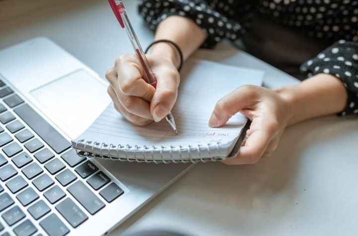 Mains qui écrivent au stylo rouge dans un carnet à spirale, à côté d’un ordinateur portable