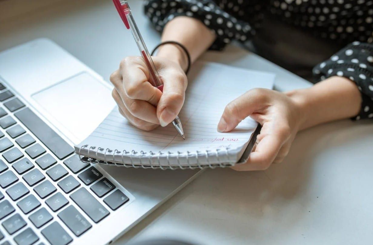 Mains qui écrivent au stylo rouge dans un carnet à spirale, à côté d’un ordinateur portable