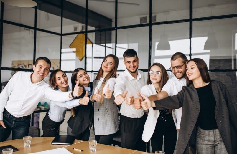 Groupe de huit jeunes adultes souriants qui lèvent le pouce dans un bureau aux cloisons vitrées