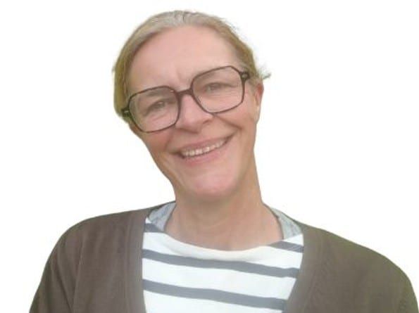 Portrait de Christine den Boer souriante, avec de grandes lunettes, un haut rayé et un gilet marron
