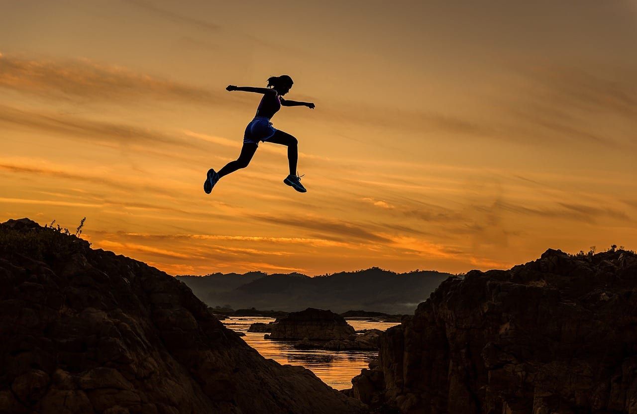 Silhouette d’une femme qui saute entre deux rochers au coucher du soleil, au-dessus d’une rivière