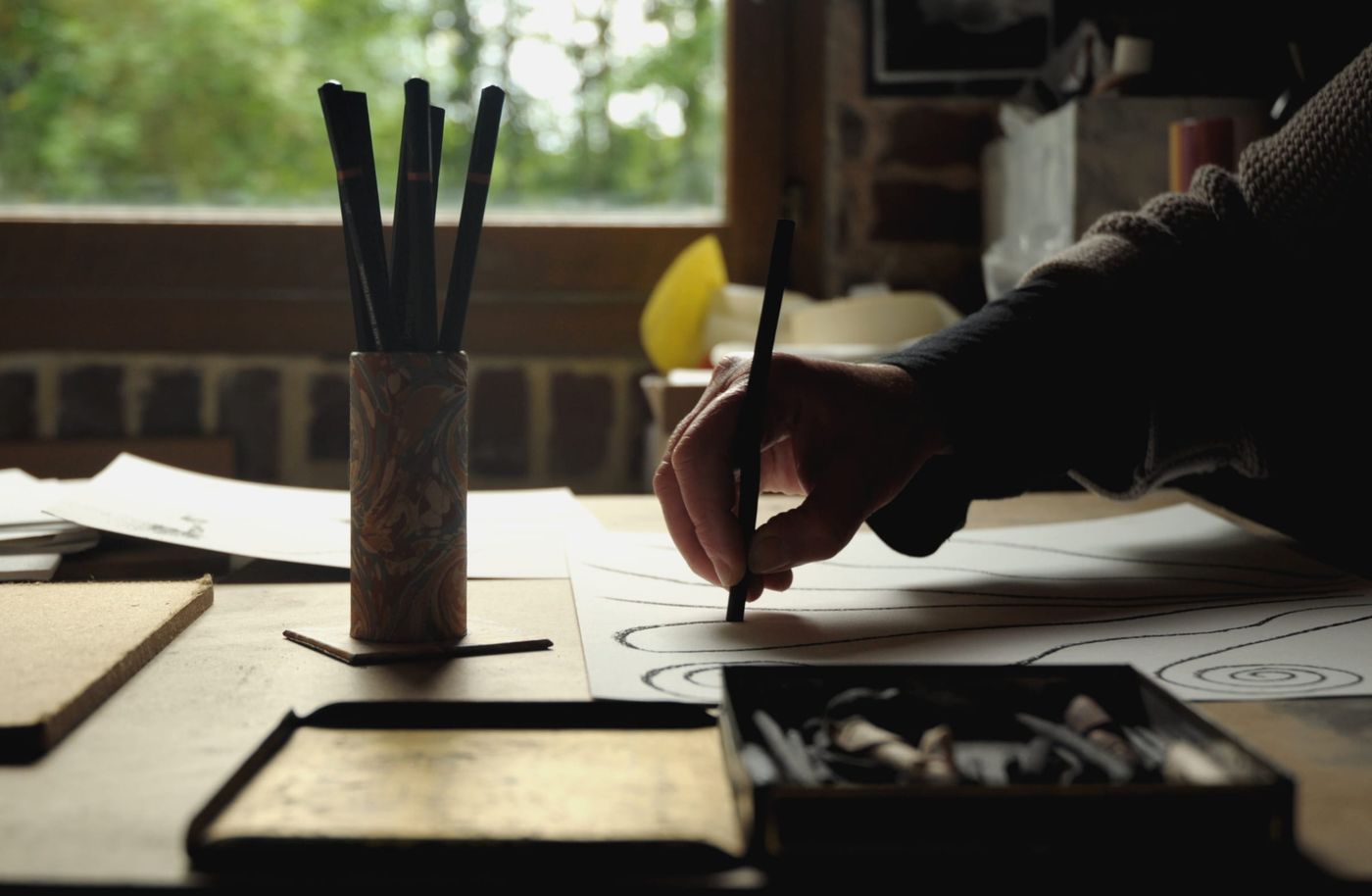 Main qui dessine des spirales au fusain sur une feuille, à côté d’un pot de crayons