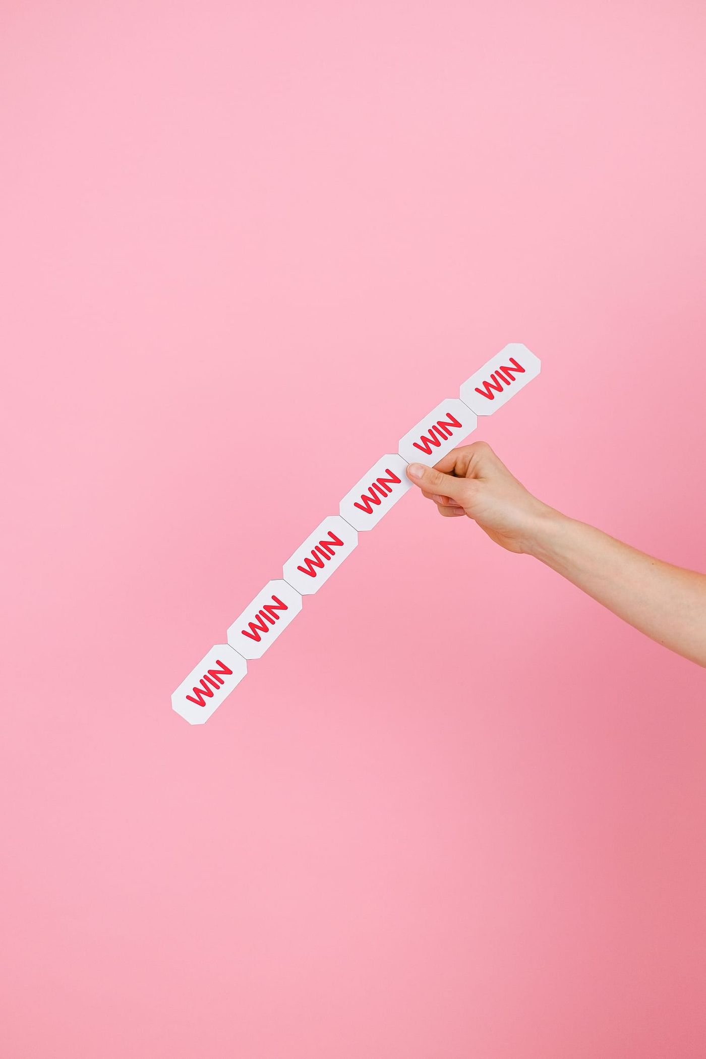 Une main tient une bande de petits tickets blancs marqués « WIN » en rouge, sur un fond rose